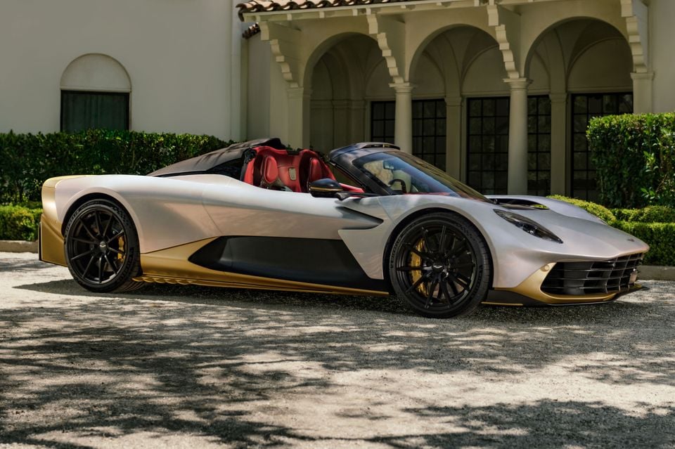 A silver and gold Aston Martin Valhalla Spider with its roof removed, parked on gravel in front of an arched villa, seen from the front side
