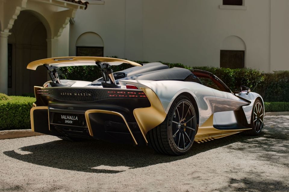 A silver and gold Aston Martin Valhalla Spider seen from the rear with its large rear wing raised, parked on gravel outside a white villa