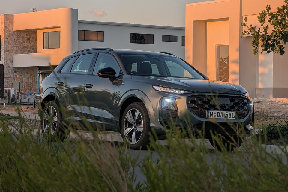 A grey Audi Q3 SUV seen from the front-side angle, parked near a modern building at sunset