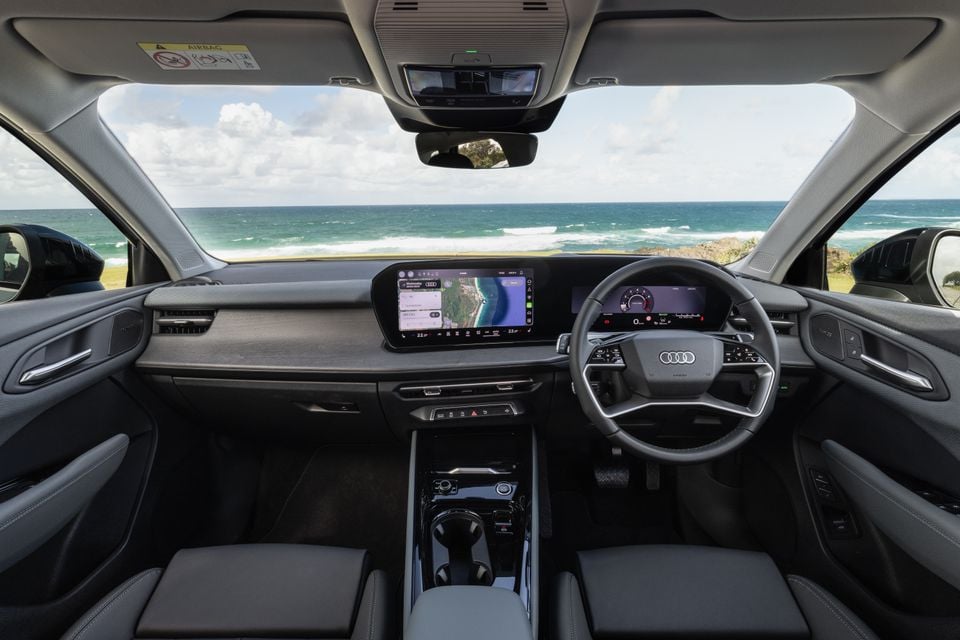 Interior view of an Audi Q3 dashboard showing dual digital screens, steering wheel with Audi logo, and centre console, with an ocean view through the windscreen