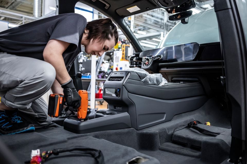 A factory worker installs the centre console inside an Audi Q9 during assembly at a manufacturing plant, using a power drill tool.