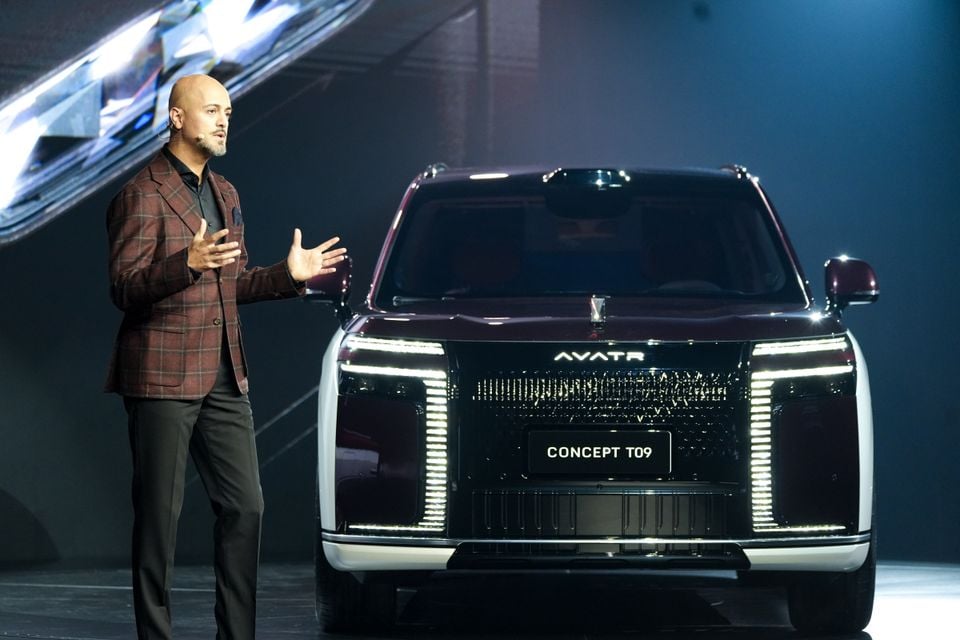 A presenter speaking on stage beside the dark red and white Avatr Concept T09 SUV, seen from the front with its illuminated headlights and grille