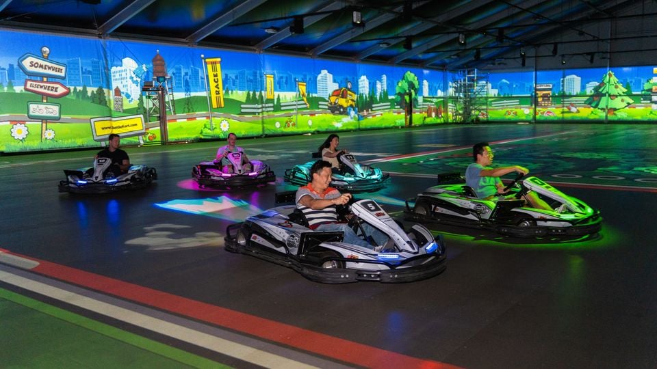 People racing colourful electric go-karts on an indoor track at BattleKart Sydney, surrounded by animated projected wall displays