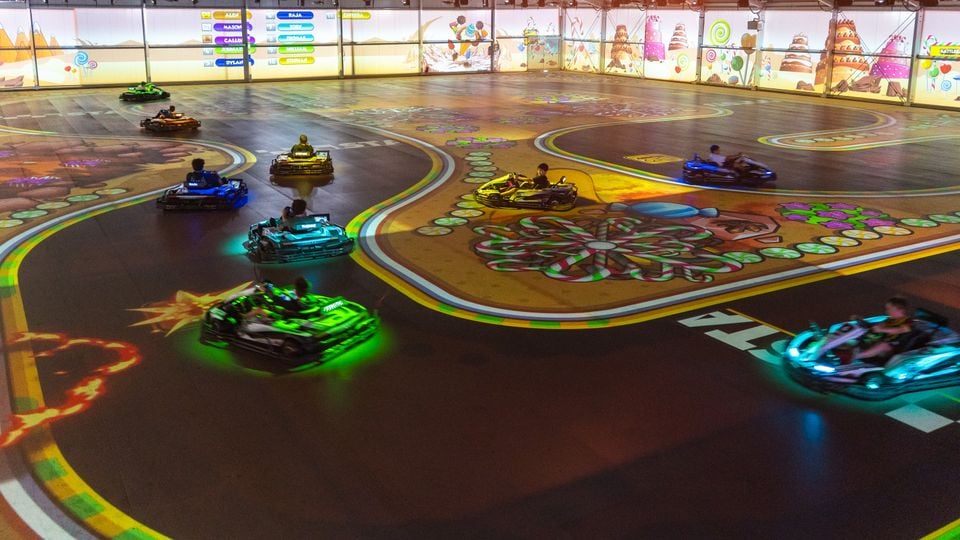 People racing colourful electric go-karts on an indoor track with projected candy-themed graphics and a scoreboard display at BattleKart Sydney