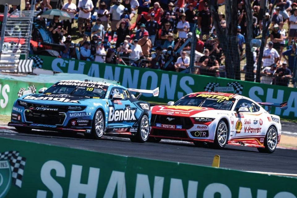 A blue Chevrolet Camaro and a red and white Shell V-Power Ford Mustang Supercar racing side by side at Mount Panorama in front of a crowd