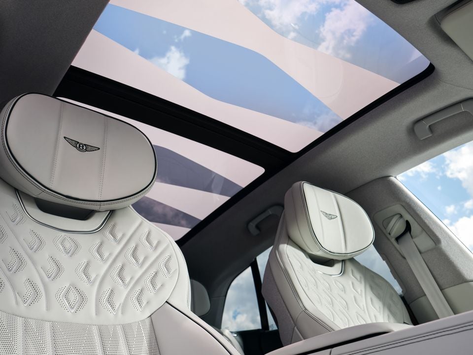 A Bentley's cream leather rear seats and quilted headrests with the Bentley wing badge, seen beneath a large panoramic sunroof showing blue sky and clouds.
