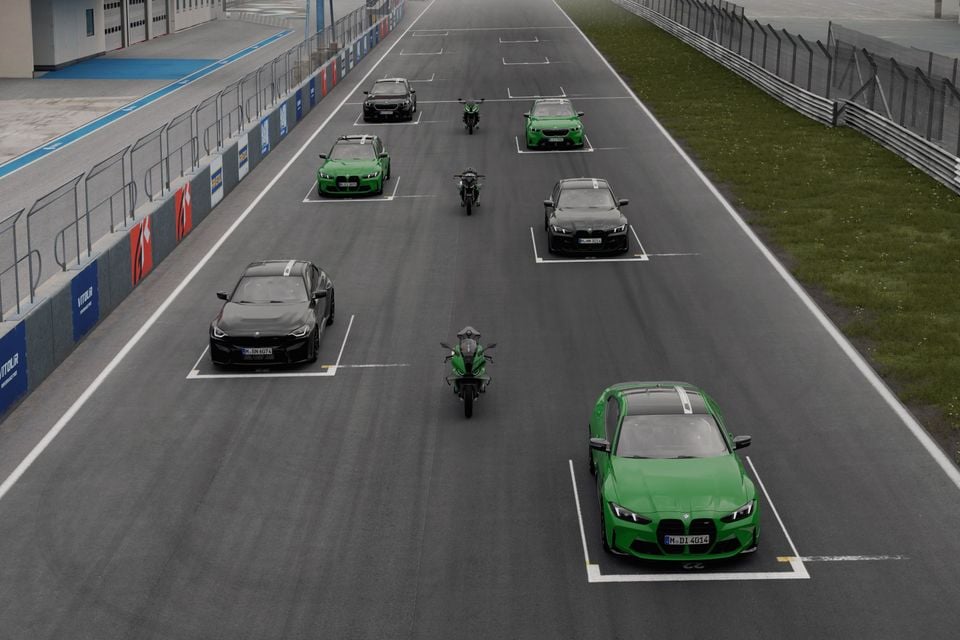 Multiple BMW cars in green and black, along with motorcycles, lined up on a motor racing circuit straight