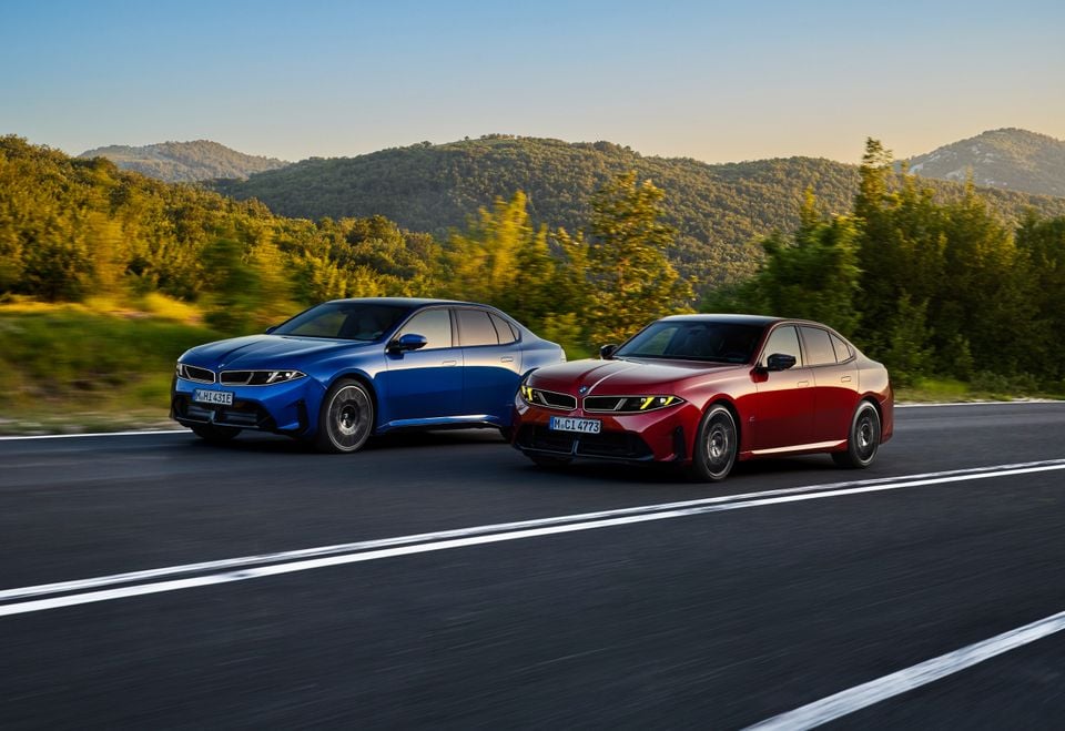 Two BMW i3 sedans, one blue and one red, driving on a road with hills in the background.