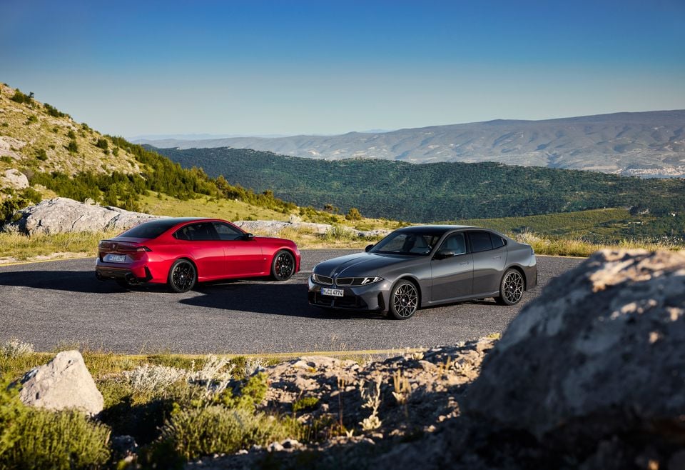 A red BMW 3 Series sedan and a grey BMW i3 sedan parked together on a mountain road overlooking a green valley.