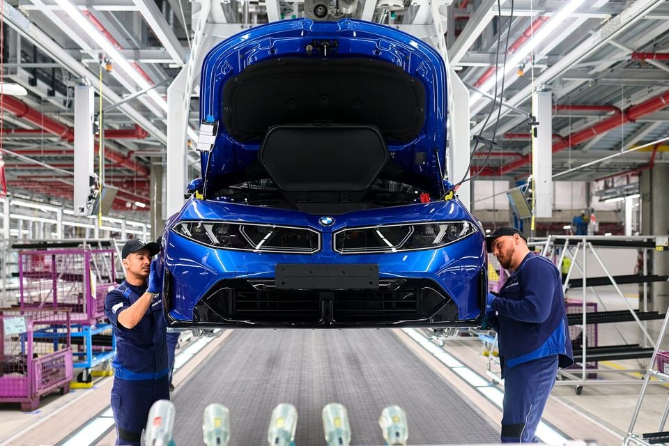 A blue BMW car is suspended on an assembly line while two factory workers inspect its front end during production.