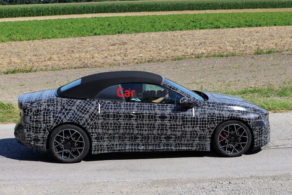 A camouflaged BMW i4 convertible prototype seen from the side on a rural road, with a soft-top roof and red brake calipers visible