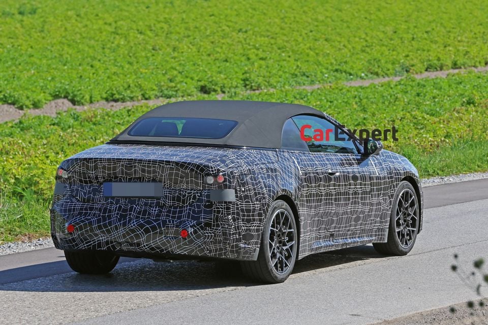 A camouflaged BMW i4 convertible prototype seen from the rear three-quarter angle on a country road