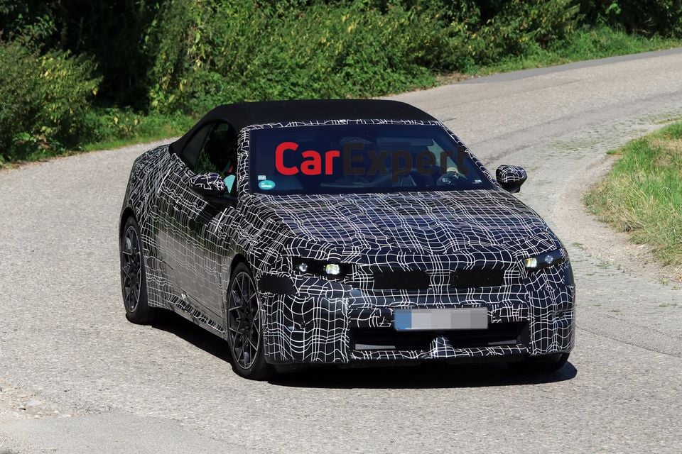 A camouflaged BMW i4 convertible prototype seen from the front on a public road during testing