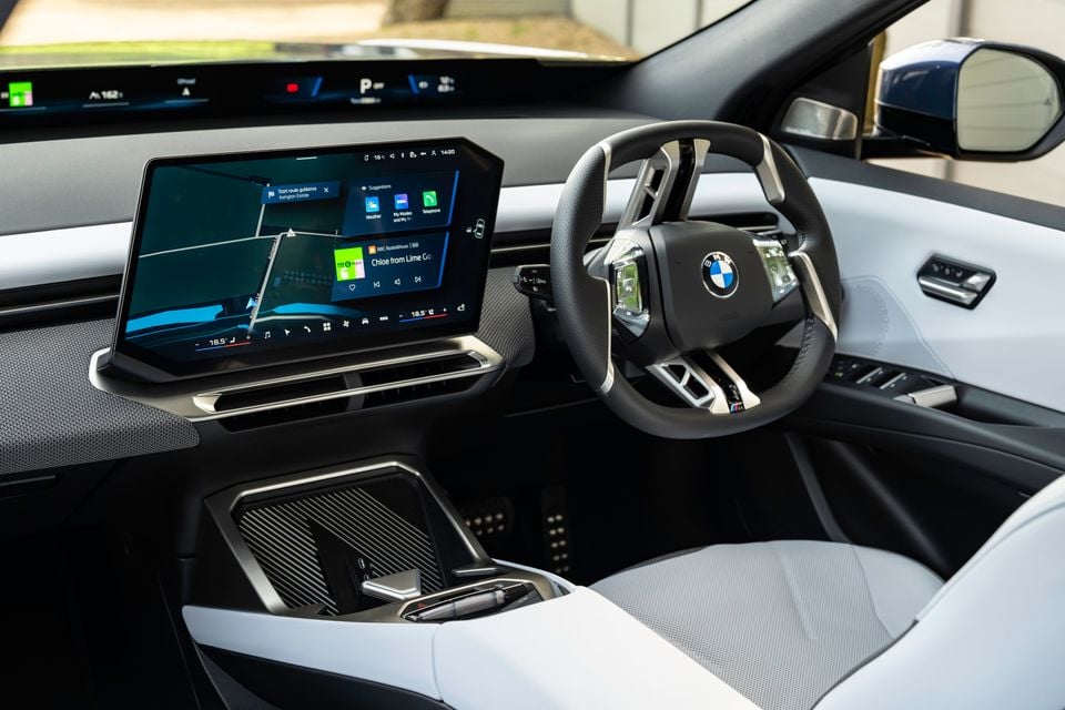 BMW iX3 interior showing the curved touchscreen infotainment display, M Sport steering wheel, digital instrument cluster, and light grey seats