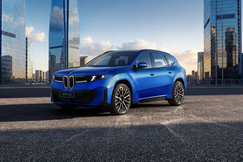 A blue BMW iX3 electric SUV seen from the front three-quarter angle, parked in an urban setting with city skyscrapers in the background.