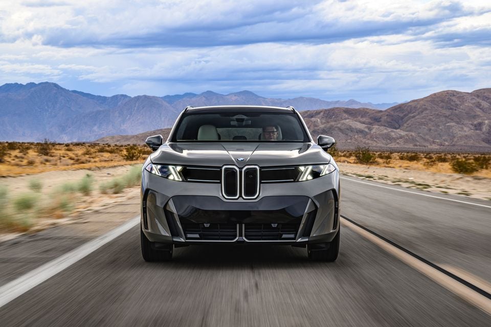 A grey BMW iX5 60 xDrive SUV driving towards the camera on a desert highway with mountains behind