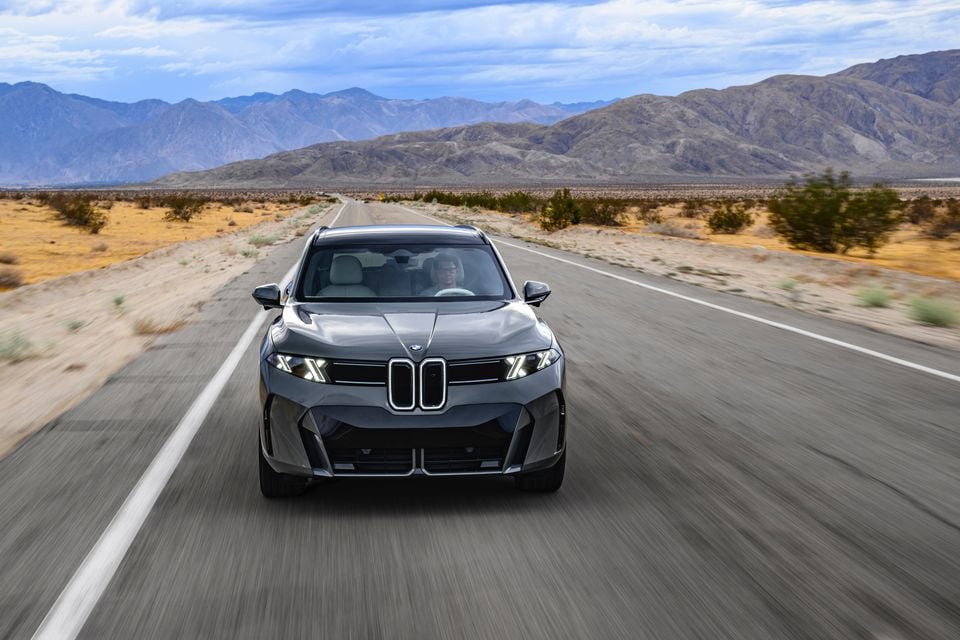 A grey BMW iX5 SUV driving towards the camera on a desert highway with mountains in the background