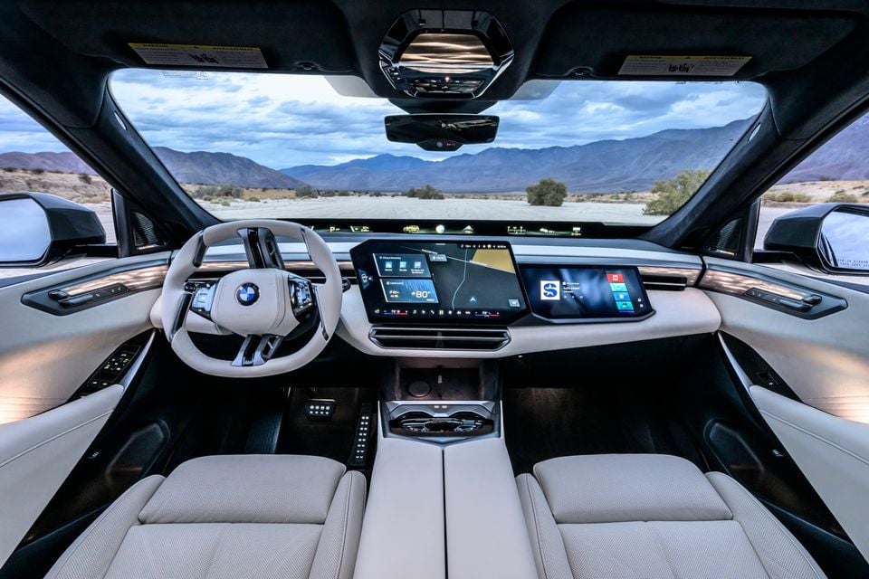 BMW cabin with a light grey interior, a minimalist steering wheel and large central touchscreen, looking out over a desert road towards mountains