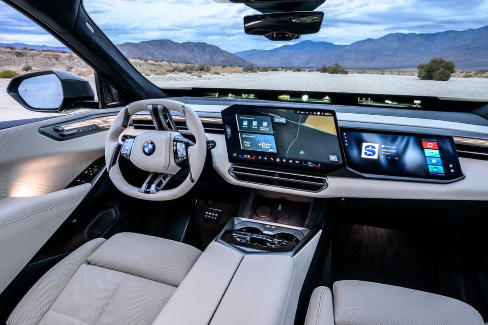 Cream leather interior of the BMW iX5 60 xDrive showing the steering wheel, curved dual touchscreens, panoramic head-up display strip and centre console