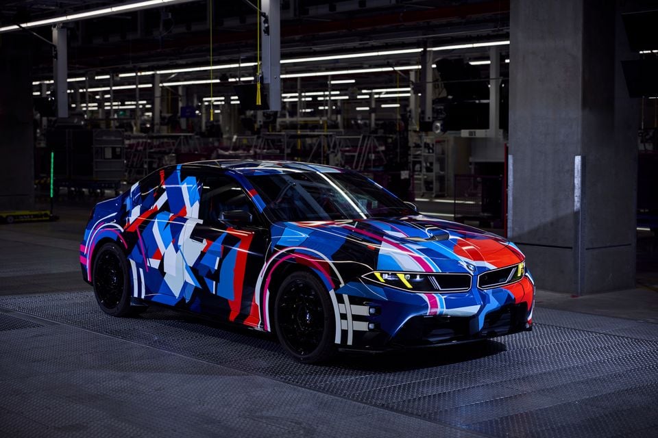 Protótipo do BMW M3 da próxima geração com camuflagem colorida em azul, vermelho e branco, estacionado dentro de uma fábrica, visto de um ângulo frontal de três quartos