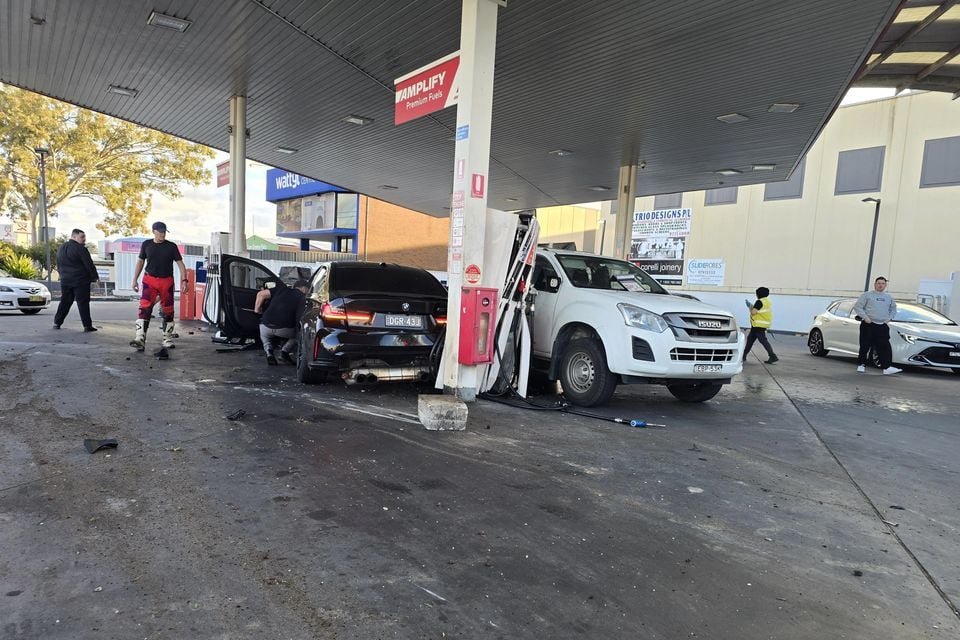 A black BMW M3 and a white Isuzu D-Max ute crashed into a petrol station bowser at an Amplify Premium Fuels service station, with debris on the ground and bystanders nearby.