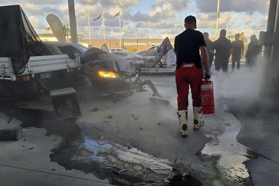 A person carrying a fire extinguisher approaches a heavily crashed car that has collided with a trailer at a petrol station, with fire suppressant foam covering the ground and smoke in the air