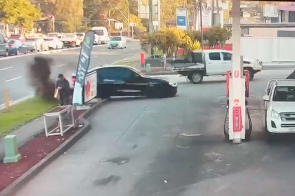 CCTV footage of a dark-coloured car crashing into a sign and kerb at a petrol station forecourt, with smoke visible and a bystander nearby