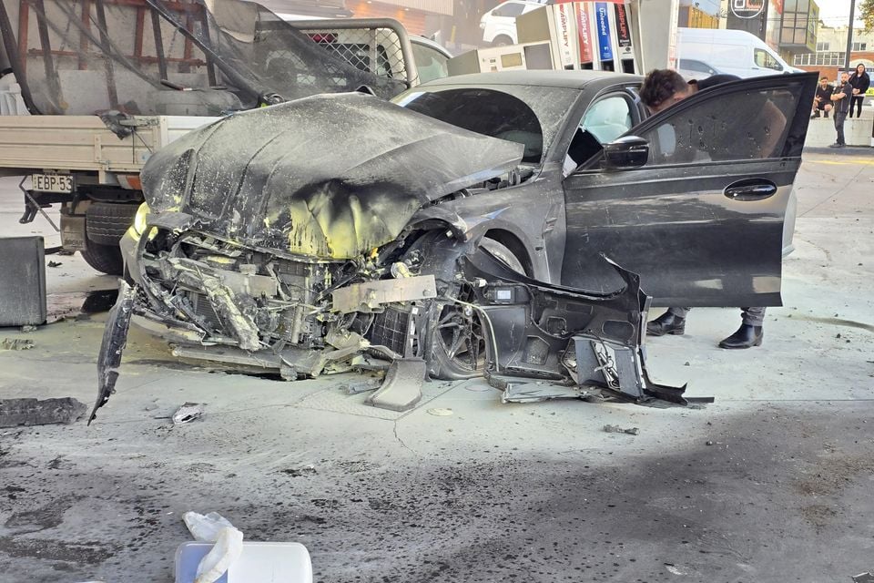 A heavily crash-damaged dark grey BMW M3 sedan with its front end completely destroyed, seen at a petrol station with bystanders nearby