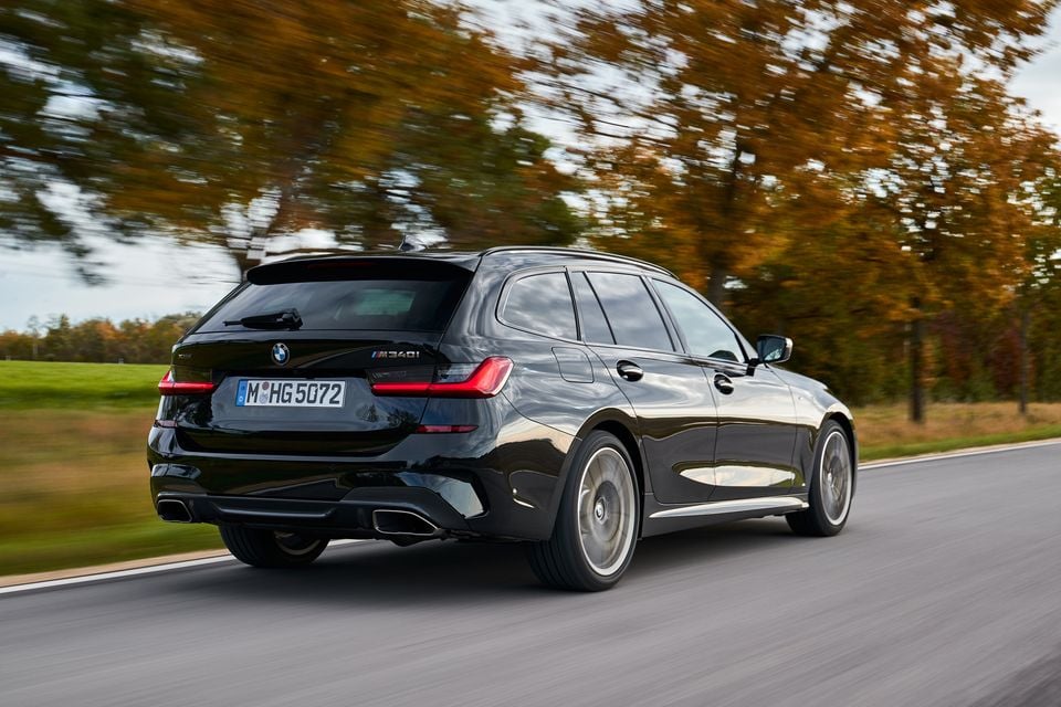 A black BMW M340i Touring wagon driving on a road, seen from the rear three-quarter angle, surrounded by autumn trees.