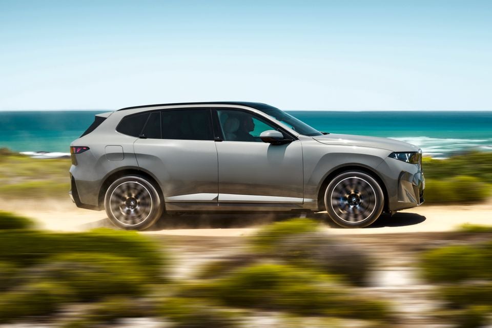 A grey BMW X5 SUV seen from the side, driving along a coastal dirt road with ocean views in the background.