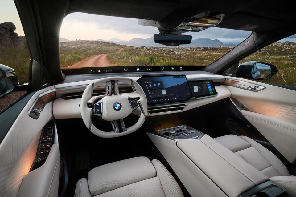 BMW X5 interior showing a large curved infotainment display, white leather seats, and a flat-bottomed steering wheel with BMW logo, with a rugged landscape visible through the windscreen