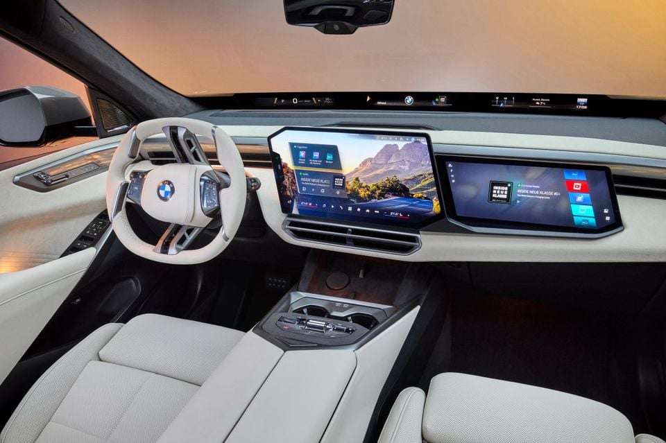 BMW X5 interior showing a large curved infotainment display, white leather steering wheel with BMW logo, cream leather seats, and a panoramic glass roof