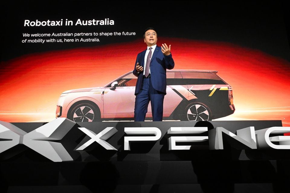 A presenter speaks on stage at an Xpeng event in Australia, with a slide behind him showing a robotaxi SUV and the text 'Robotaxi in Australia'