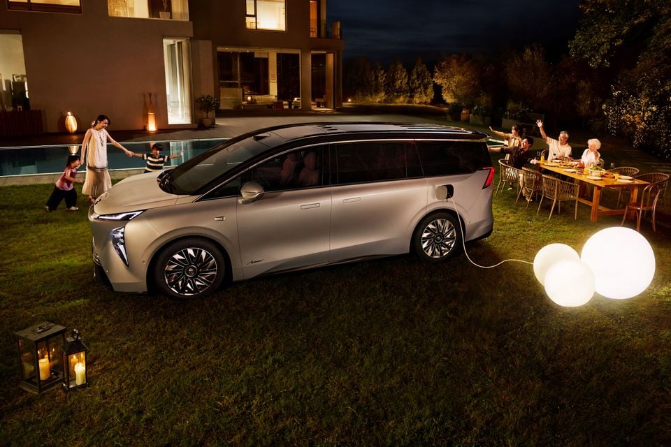 A silver Buick Electra Encasa electric minivan parked on a lawn at night, plugged in and powering outdoor globe lights, with a family gathered nearby