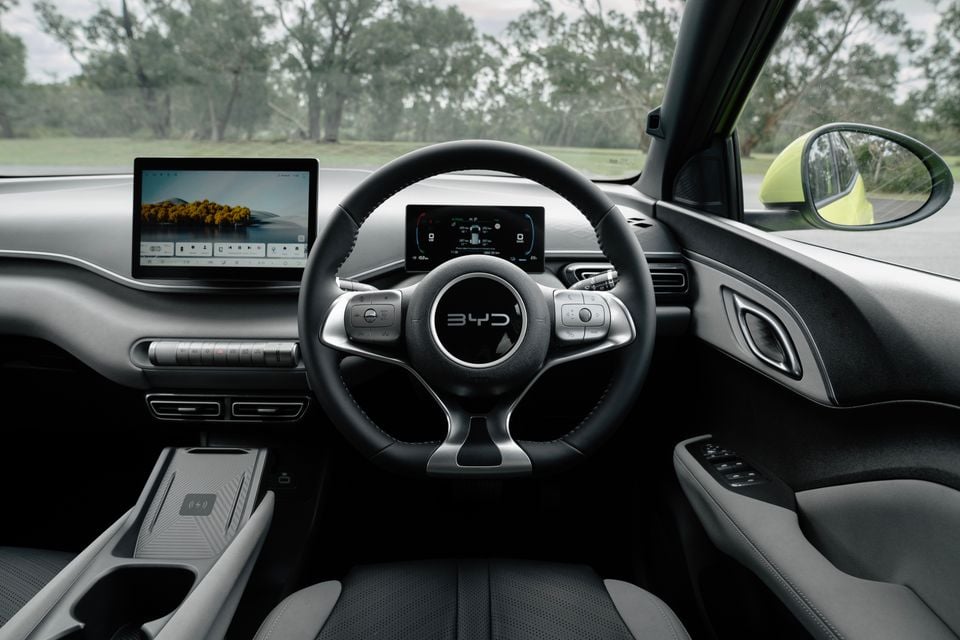 Interior of a BYD Atto 1 showing the steering wheel with BYD logo, digital instrument cluster, large central touchscreen infotainment system, and wireless charging pad, shot from the driver's seat in right-hand drive configuration