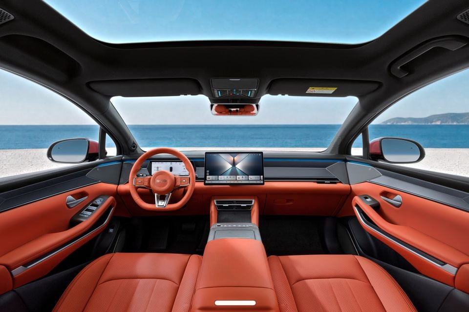 Orange leather interior of the BYD Atto 8 with steering wheel, digital driver display, central touchscreen and panoramic glass roof overlooking the sea