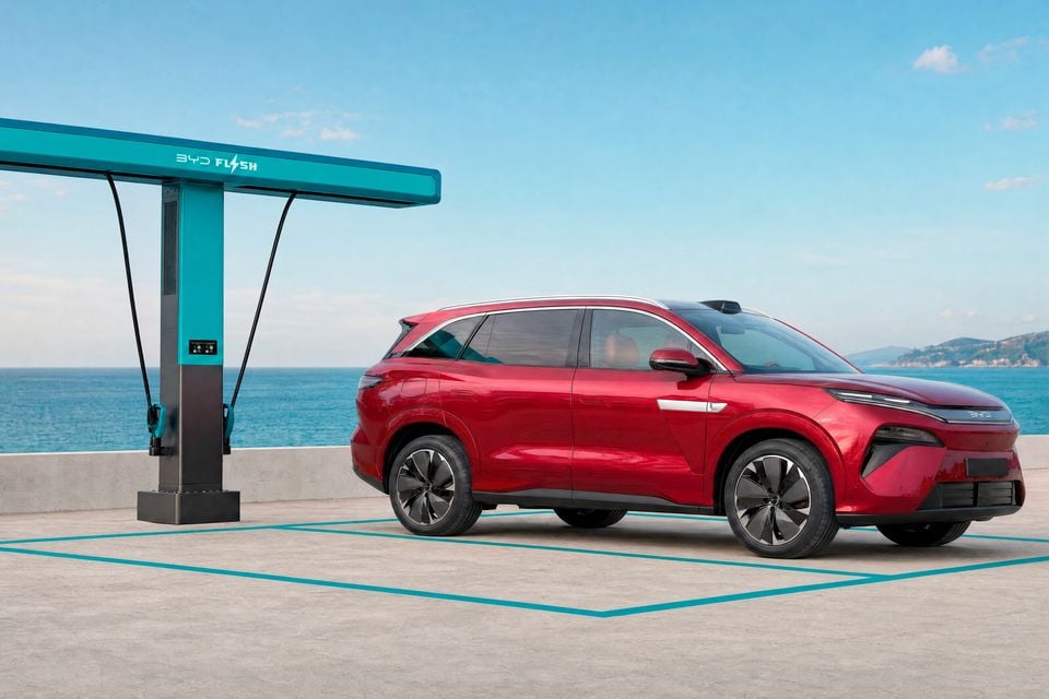 A red BYD Atto 8 SUV seen from the front three-quarter angle, parked beside a BYD Flash charging station overlooking the sea