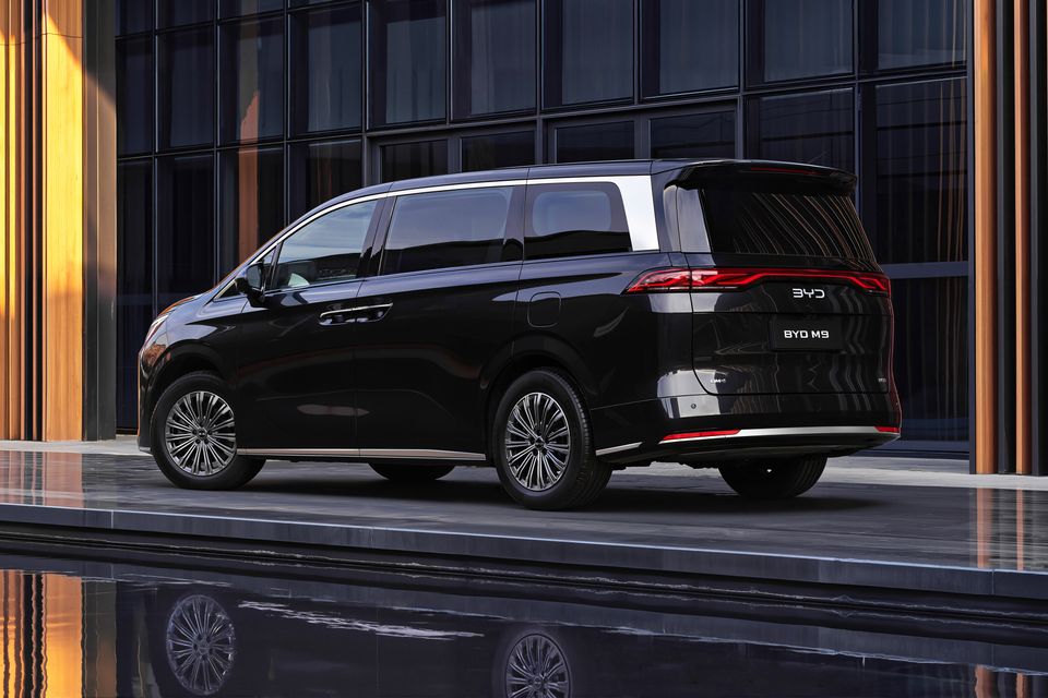 A black BYD M9 minivan seen from the rear three-quarter angle, parked in front of a modern glass building