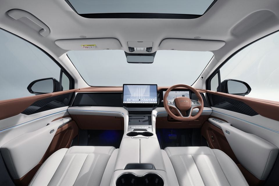 Interior of a BYD M9 showing the white and tan leather cabin with a large central touchscreen, right-hand-drive steering wheel, panoramic sunroof, and ambient blue lighting