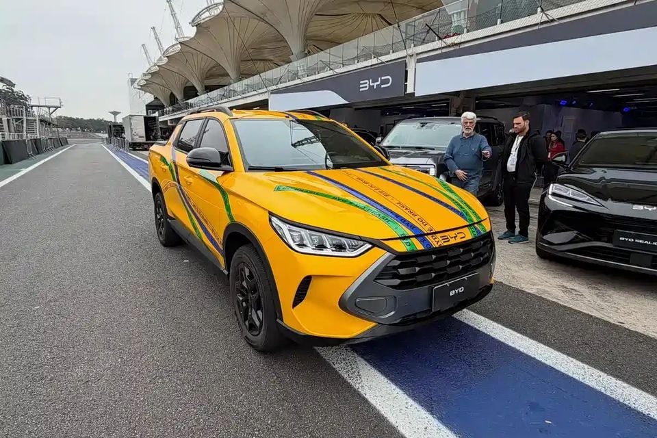 A yellow BYD Mako ute with Brazilian-themed green and blue stripe livery on the bonnet and sides, parked in a motorsport circuit pit lane with a BYD branded garage and a BYD Seal U visible in the background