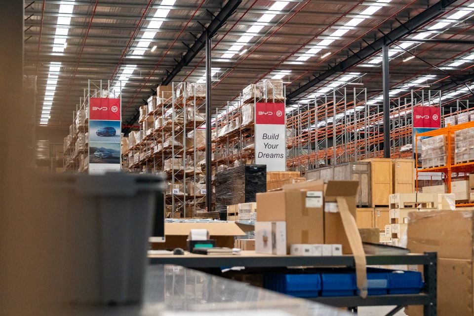 Inside the BYD and SF Express parts warehouse, with tall racks of boxed stock, BYD banners and a packing bench in the foreground
