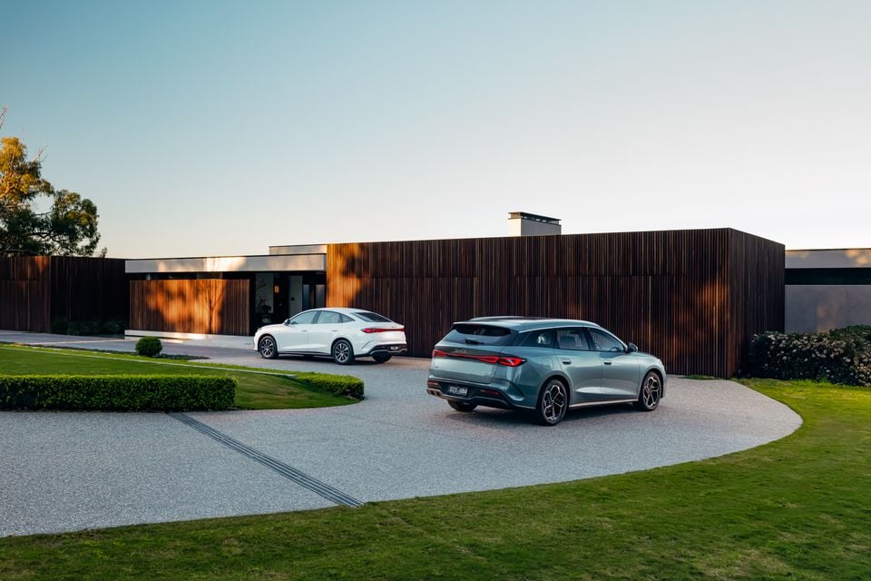A grey BYD SUV wagon and a white BYD sedan seen from the rear, parked on a gravel driveway in front of a modern timber-clad home at dusk