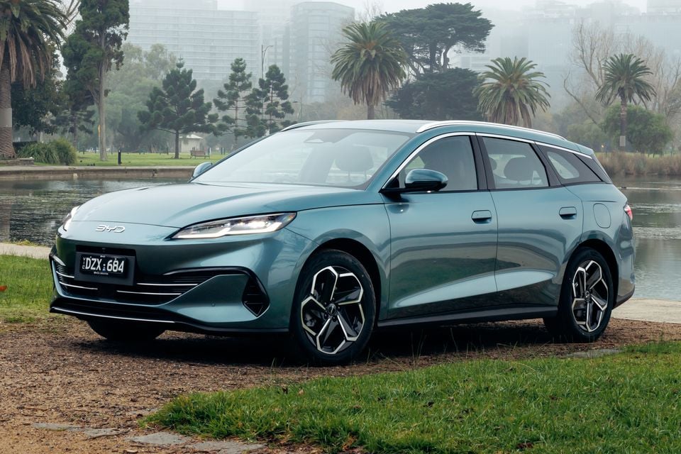 A teal BYD Seal 6 Touring wagon parked on grass beside a lake, seen from the front left with city buildings in the misty background