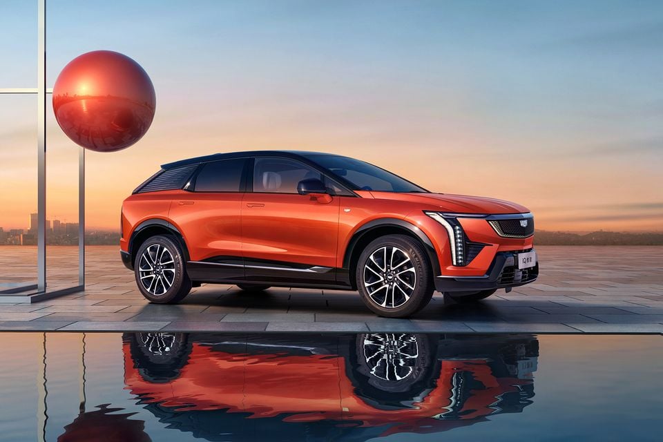 An orange Cadillac Optiq SUV seen from the front-side angle, reflected in a pool of water at dusk with a red spherical sculpture in the background.