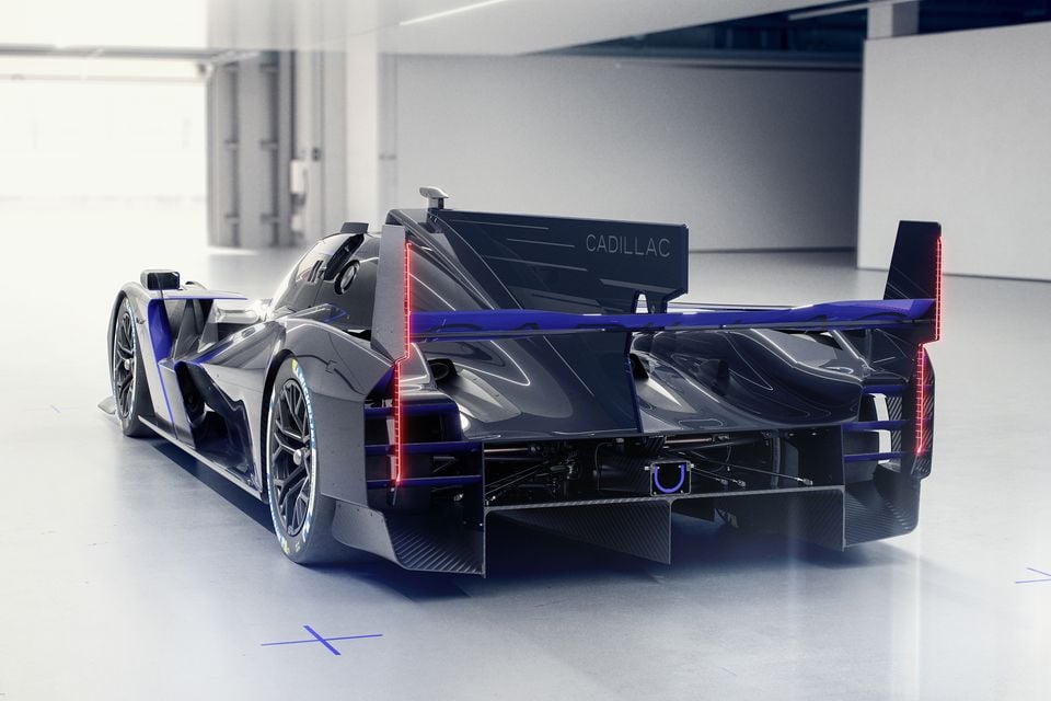 The rear of the Cadillac V-LMDh race car concept, showing its large rear wing, red LED tail lights, blue accents, and exposed carbon fibre bodywork in a studio setting.