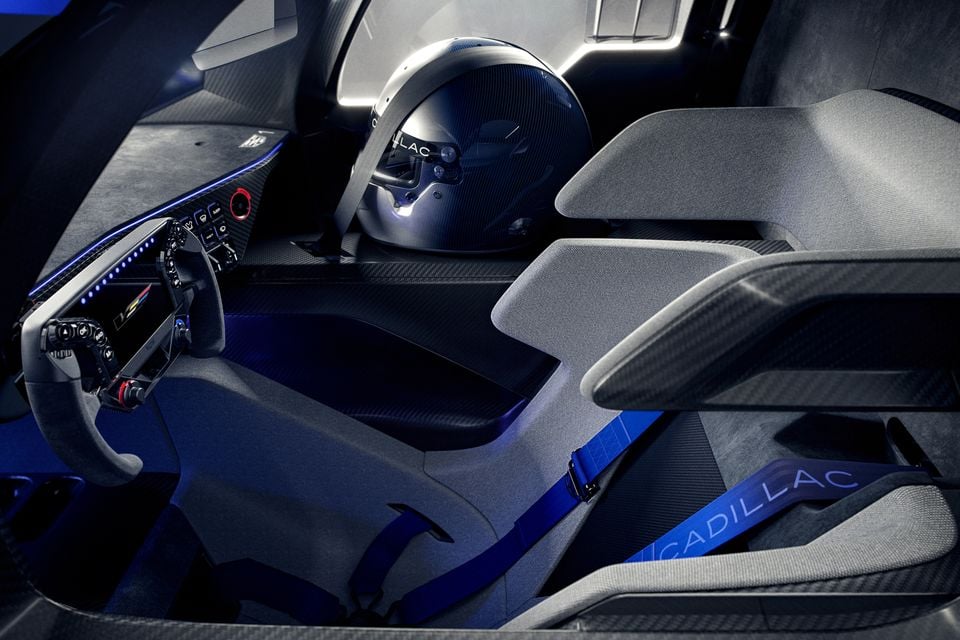 Interior of the Cadillac V-One concept showing racing bucket seats with blue Cadillac-branded harness belts, carbon fibre trim, a flat-bottomed steering wheel, digital dashboard, and a Cadillac racing helmet resting on the passenger seat