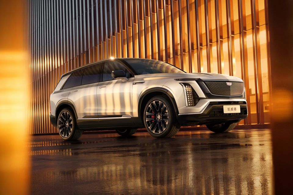 A silver Cadillac Vistiq large SUV seen from the front-side angle, parked against a warmly lit architectural backdrop