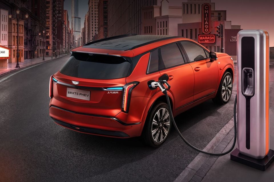 A red Cadillac XT5 PHEV SUV plugged into a charging station, seen from the rear three-quarter angle on a city street.