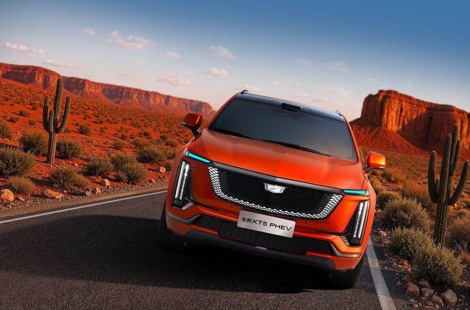 An orange Cadillac XT5 PHEV SUV seen from the front on a desert road with red rock formations and cacti in the background.