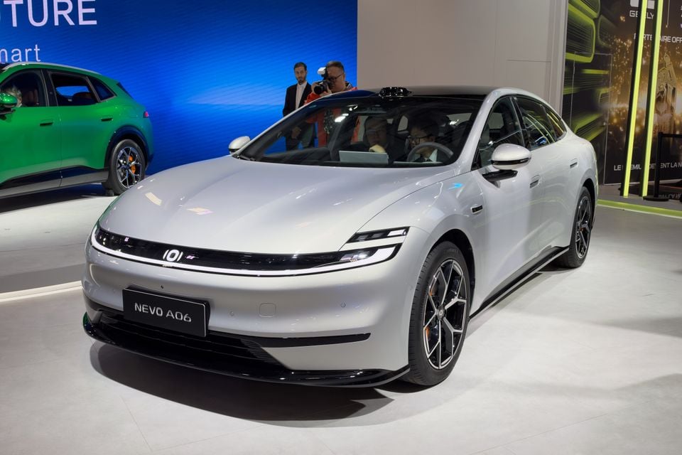 A silver Changan Nevo A06 sedan seen from the front on display at a motor show, with people seated inside and a photographer behind